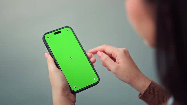 Over shoulder view close up hand of Asian woman holding smartphone green screen mockup with tracking markers, female tapping mobile phone Chroma key display outdoors for digital app design, technology