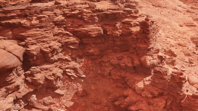 Red rock formations fill the landscape in a dry area under bright sunlight. Shadows highlight the textures and shapes of the rocks. The scene shows a barren environment, typical of desert regions.