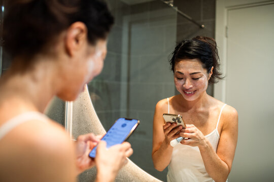 Smiling woman with face mask texting in home bathroom mirror