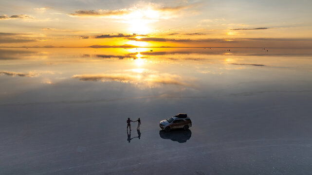 Aerial view of Uyuni Salt Flat at sunset with a couple holding hands and an SUV reflected in the shallow water, Potosi Department, Bolivia.