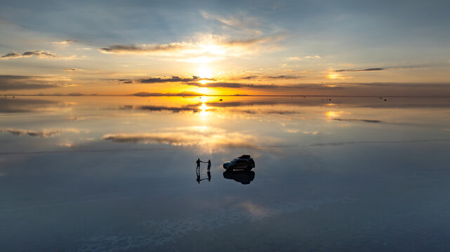 Aerial view of Uyuni Salt Flat at sunset with a couple standing near their parked vehicle, creating reflections on the water surface in Potosi Department, Bolivia.