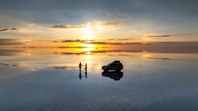 Aerial view of Uyuni Salt Flat at sunset with a couple standing and holding hands near their parked car, creating perfect reflections on the water surface in Potosi Department, Bolivia.