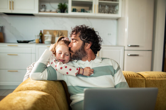 Father kissing daughter on sofa at home with laptop