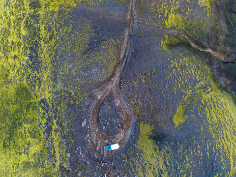 Aerial view of a dirt road loop winding through a vibrant green mossy volcanic landscape with a small vehicle parked at the edge in Iceland.