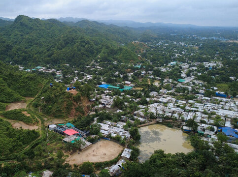 Aerial view of the Rohingya refugee camp with dense rows of shelters, a small pond, and surrounding lush green hills in Cox's Bazar, Chittagong Division, Bangladesh.