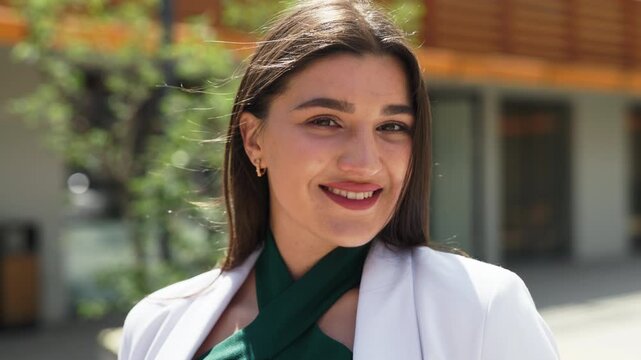 Charming businesswoman in white blazer posing outside in sunny weather