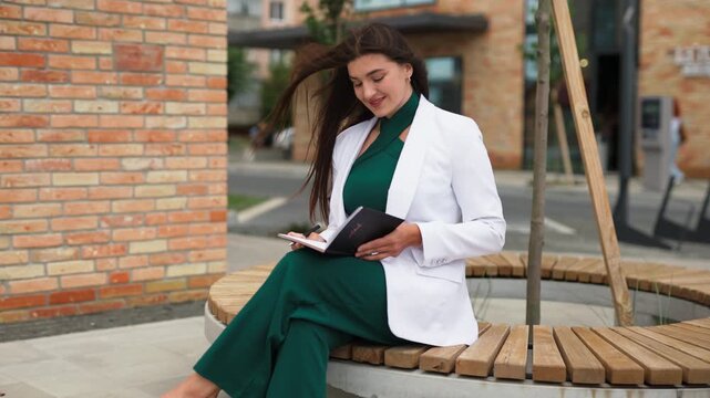 Beautiful young business woman sitting on bench and writing in notebook outdoors, slow motion