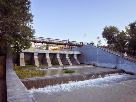 Urban Flood Control Dam with Pedestrian Bridge and Flowing River at SunsetA modern flood control dam with cascading water, a pedestrian bridge, and riverside infrastructure surrounded by lush trees un