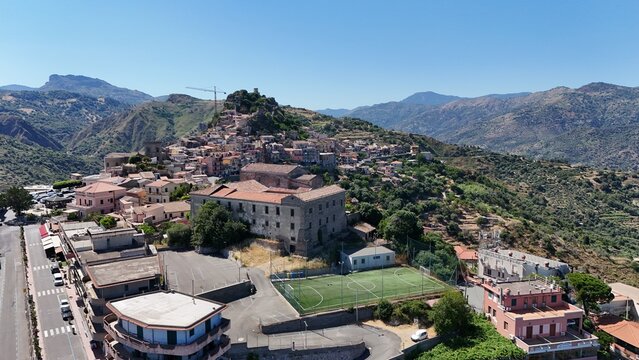 Aerial view of the historic hilltop village of Forza d'Agro, featuring stone buildings, a soccer field, and surrounding mountainous landscape in Forza d'Agro, Sicilia, Italy.