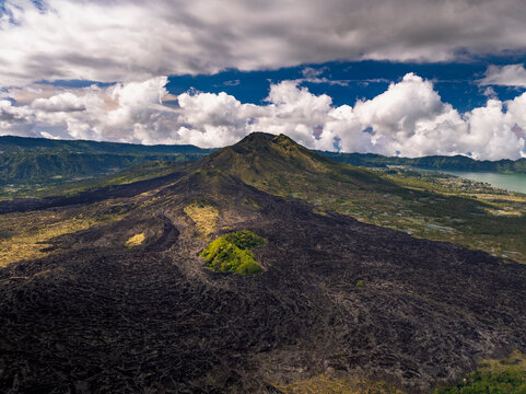 Aerial view of Mount Batur volcano featuring expansive black lava fields, a patch of lush green vegetation, and Lake Batur under a dramatic cloudy sky in Kintamani, Bali, Indonesia.