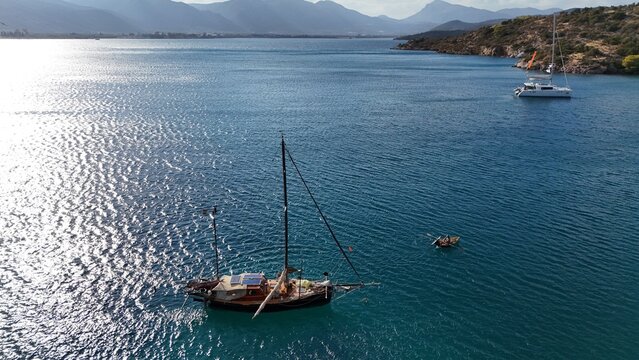 Aerial view of a sailboat anchored in the clear blue waters of the bay, with a small rowing boat nearby and a catamaran in the distance, Poros, Greece.