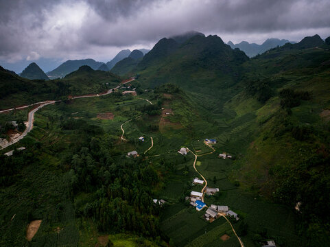 Aerial view of the winding roads of the Ha Giang Loop cutting through lush green mountains and small rural settlements under a cloudy sky in Ha Giang, Ha Giang, Vietnam.