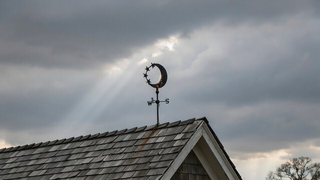 Metal weather vane with a crescent moon and star motif, perched on a shingled roof with a sunbeam breaking through dark, heavy storm clouds