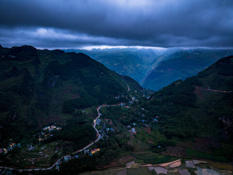 Aerial view of Dong Van town nestled in a deep valley surrounded by dramatic mountains, winding roads, and terraced fields under a moody twilight sky in Dong Van, Ha Nam, Vietnam.