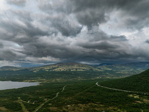 Aerial view of the mountainous landscape near Dombas with a winding road, a lake, and dramatic dark storm clouds over the hills in Dombas, Innlandet, Norway.