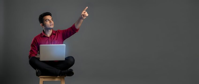 Young Asian man in burgundy sweater sitting cross-legged with laptop pointing upward, expressing eureka moment or breakthrough idea for business presentations.