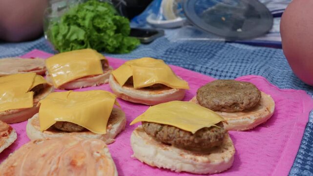 Fresh hamburger buns are arranged in a row with melted cheese and beef patties during outdoor preparation. A bowl of lettuce and food supplies rest on a blue picnic blanket in the background.