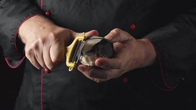 Chef in black uniform with red accents peels a round vegetable using a yellow peeler, showcasing the continuous action of removing the skin in a well-lit kitchen setting