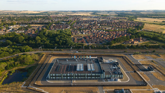 Aerial view of a hyperscale data center during golden hour, featuring industrial rooftops, cooling units, and perimeter fencing, near a residential area in Didcot, England, United Kingdom.