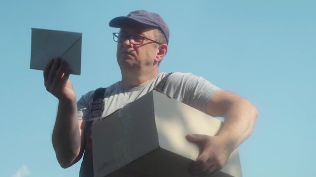 A male postman in a gray shirt and blue cap examines a letter while holding a large cardboard box outdoors, against a clear blue sky, illustrating the process of handling mail and parcels
