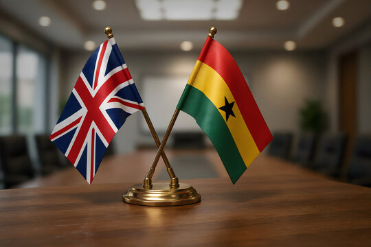 Two small desk flags of the united kingdom and ghana crossed on a wooden table in a blurred office conference room setting for diplomacy