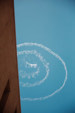 Spiral white smoke contrails or skywriting against a clear blue sky, framed by a concrete building wall