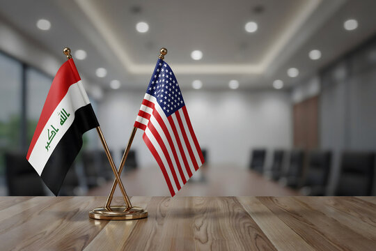 Crossed miniature flags of iraq and the united states on a wooden conference table in a blurred office room with soft overhead lighting fixtures