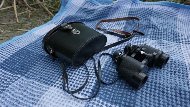 Black vintage binoculars and a leather carrying case lie on a blue patterned picnic blanket. The optical equipment is placed outdoors on the grass during a scenic adventure trip.
