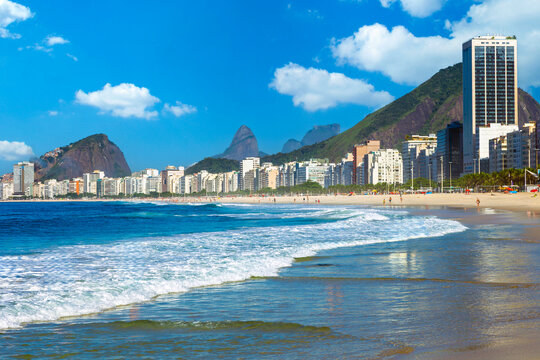 Beautiful sunny day at Copacabana beach in Rio de Janeiro, Brazil, featuring ocean waves, city skyline, and the iconic Dois Irmaos and Pedra da Gavea mountains in the background.
