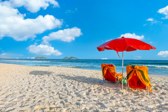 Red umbrella and two beach chairs on the white sands of Barra da Tijuca beach, Rio de Janeiro. A perfect sunny summer day with blue sky and ocean views.