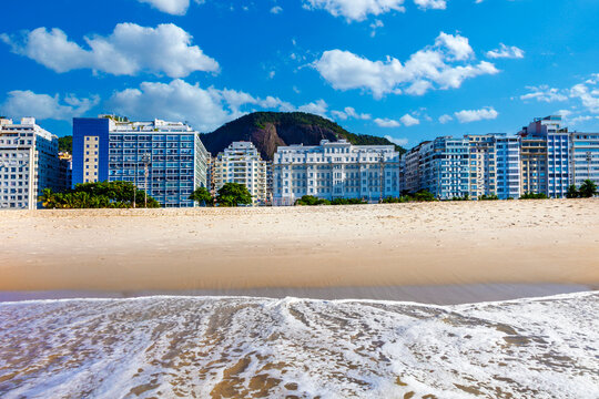 Scenic view of Copacabana Beach in Rio de Janeiro, Brazil. White ocean foam meets the sand, with a long line of hotels, including the Copacabana Palace, and mountains under a cloudy blue sky.