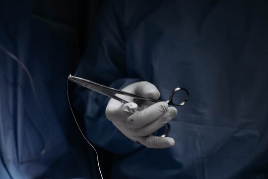 Gloved surgeon holding a surgical needle holder and suture during an operation, showing precise suturing technique and sterile manual work in a modern surgical environment. Medical concept.