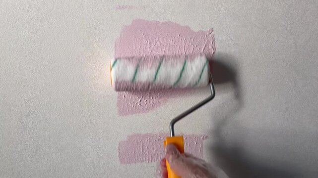Close-up of a hand painting a textured wall with light pink paint using a roller. A home renovation, DIY repair, or interior decoration process.