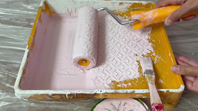 Close-up of a paint roller being loaded with light pink paint in a yellow tray