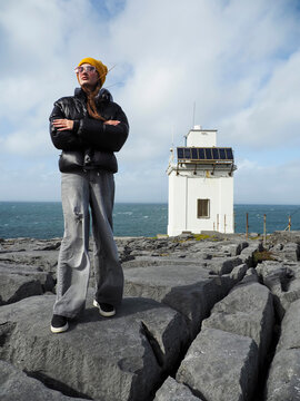 Girl tourist in black jacket and yellow hat is standing on rough stone surface, Blackrock lighthouse in the background. Burren area of Ireland. Popular tourist location and point of interest. Travel.