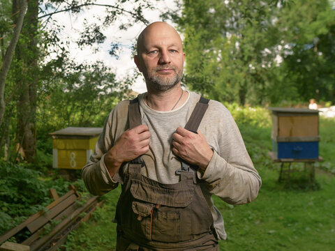 Bald man in dirty overalls stands near beehives. Trees and green grass surround him. He looks at camera with calm expression. Rural farmland background. Male in his 40s or 50s. Farmer portrait.