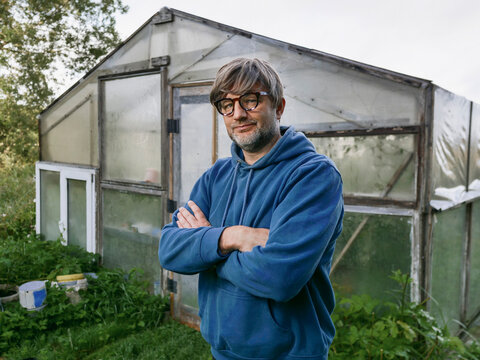 Man stands in wooden greenhouse with glass panels. He wears blue hoodie and glasses and he is in his 40s or 50s with grey beard and hair. Hands on hips, looks at camera. Growing organic produce.
