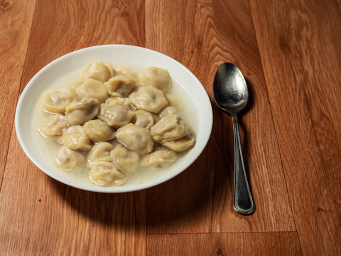White bowl filled with dumplings in broth, placed on wooden table. Metal spoon rests beside the bowl. Simple, rustic meal presentation with warm lighting. Popular Russian dish. Fast food. Simple meal.