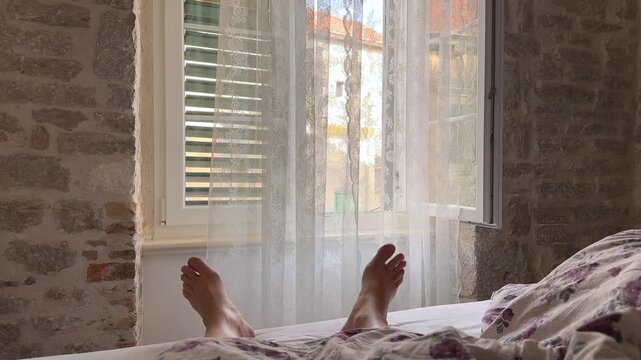 Person relaxing in bed with feet visible against a window with white curtains and stone walls