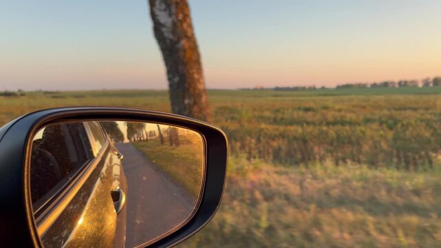a car's side wing mirror reflecting a narrow paved road lined with trees as the vehicle drives through the countryside.