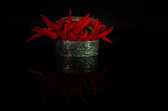 Red hot chili peppers displayed in a decorative silver bowl against a black background. Minimal food composition with glossy reflections, bold color contrast and copy space for culinary concepts.