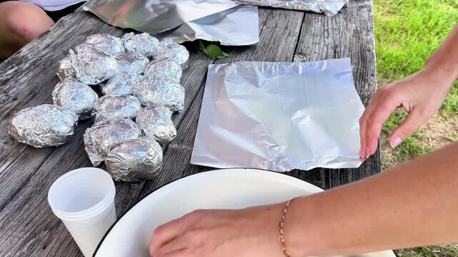 Woman's hands wrapping fresh potatoes in silver aluminum foil sheet on a wooden picnic table outdoors to prepare them for grilling over a bonfire campfire.