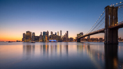 Naklejka premium Panoramic sunset view of the New York City skyline with the Brooklyn Bridge illuminated over the East River during twilight hour with city reflections
