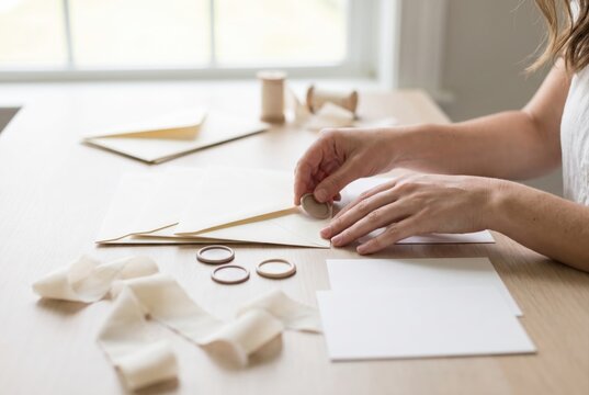 Hands pressing a beige wax seal onto the flap of a blank paper envelope on a wooden desk
