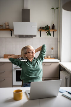 Dreamy happy middle aged woman sits back in chair with satisfied expression, resting stretching arms after finishing long working day and tasks. Female enjoying working on remote from cozy kitchen.