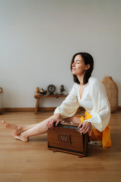 Woman sits barefoot with harmonium indoors, photographic, on a warm wooden floor, symbolizing mindfulness and wellness