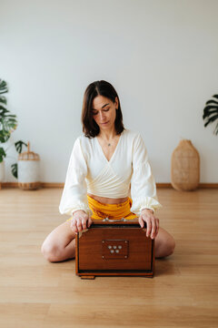 Photographic woman in white blouse sitting with harmonium in a calm, plant-filled studio, expressing mindful meditation