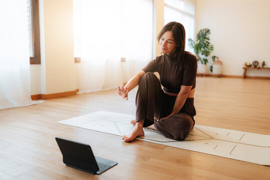 Woman practicing yoga indoors in a lifestyle photograph, seated on a mat with a laptop on a sunlit wooden floor, wellness concept
