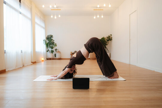 Photographic woman practices downward dog yoga on a mat during an online workout in a home with plants and warm wood floor