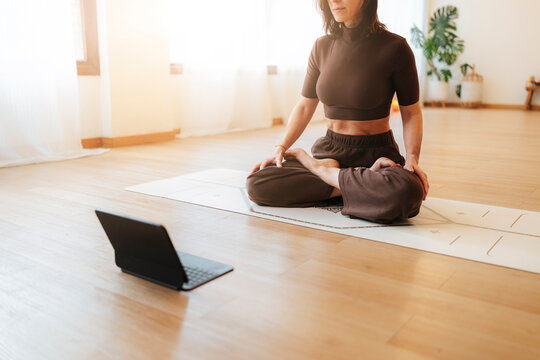 Woman practices seated yoga on a mat beside a laptop in a sunlit studio, warm photographic style, wellness and mindful exercise